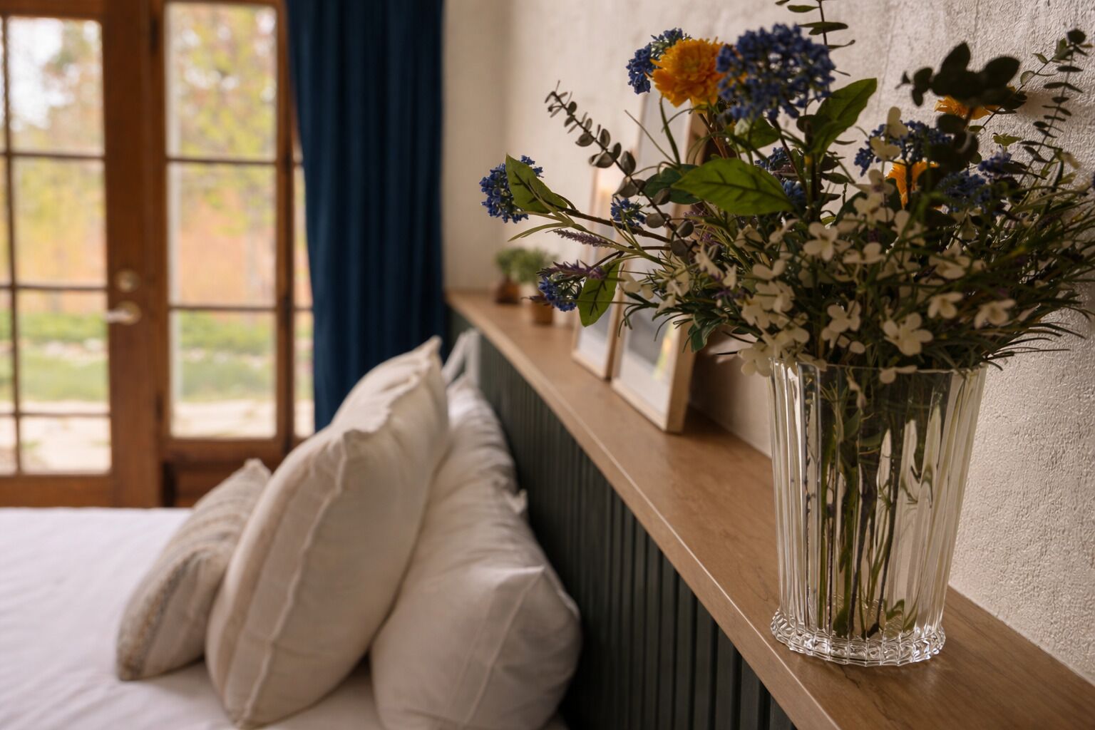 A bedroom with a bed, white pillows, a bedside table with a vase of flowers, and a view through a window.