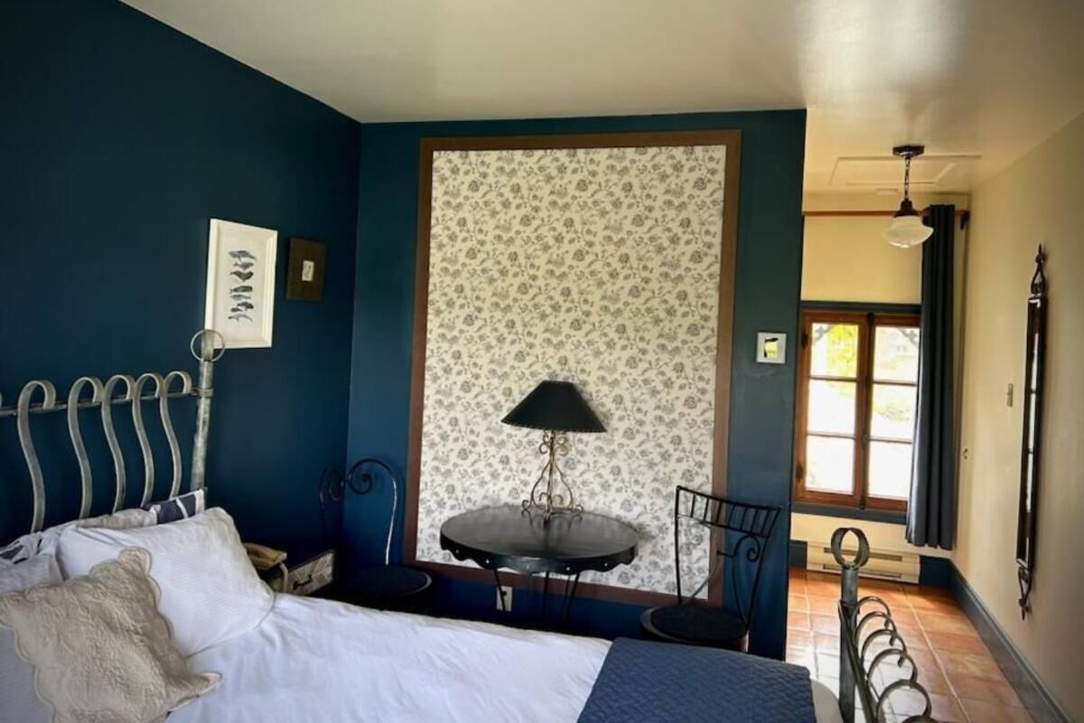 Une chambre à coucher avec un lit, une petite table avec une lampe, une chaise et une fenêtre avec des rideaux.