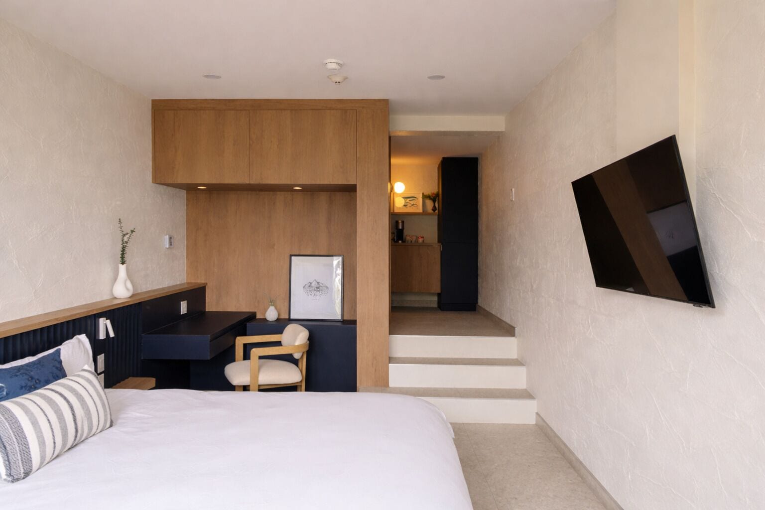 A modern hotel room with a bed, a desk, a chair, a TV, and a kitchen area in the background.