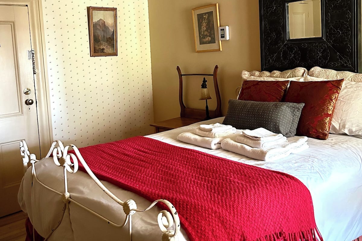 Un lit bien fait, recouvert d’une couverture rouge, une table de chevet avec une lampe, un miroir fixé au mur et une porte ornée d’un papier peint à motifs.