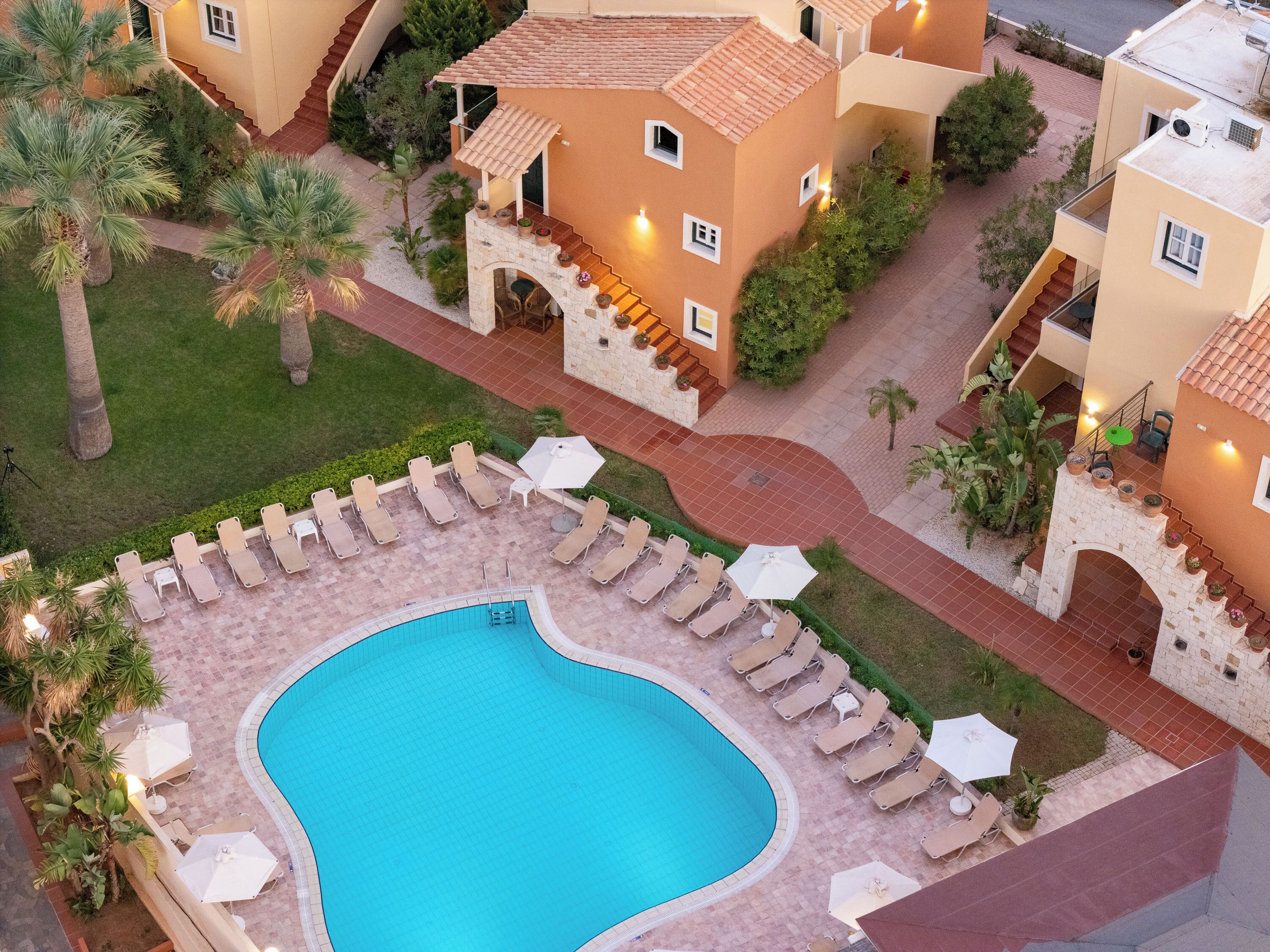 A pool area with lounge chairs and umbrellas, surrounded by a garden with palm trees and a stone pathway.