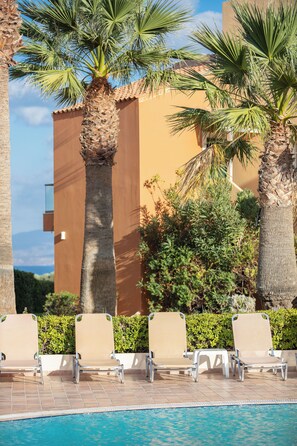 A poolside area with lounge chairs, palm trees, and a building in the background. - Dia Apartments (Hersonissos)