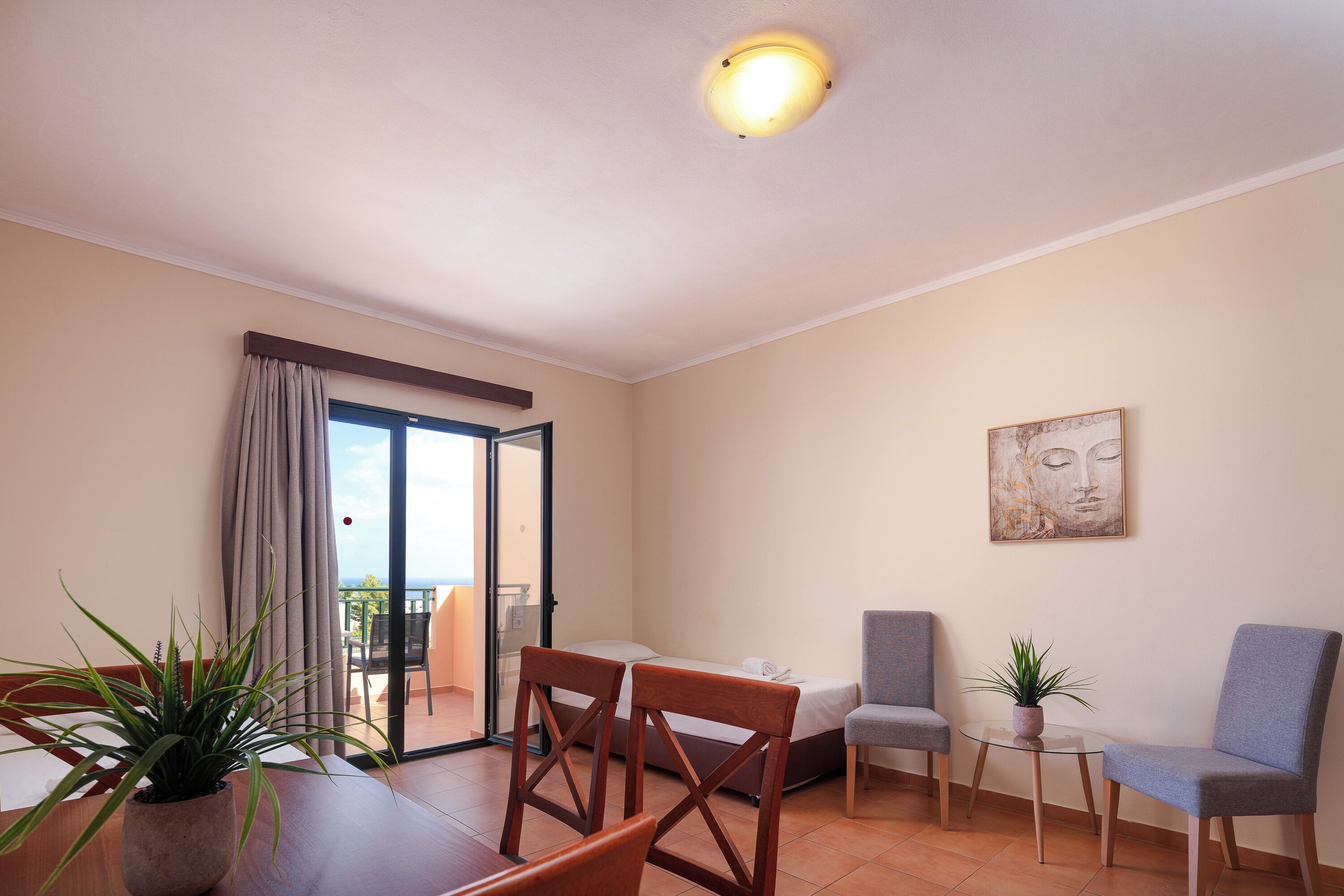 A hotel room with a bed, two chairs, a small table, a potted plant, a painting, and a balcony with a view.