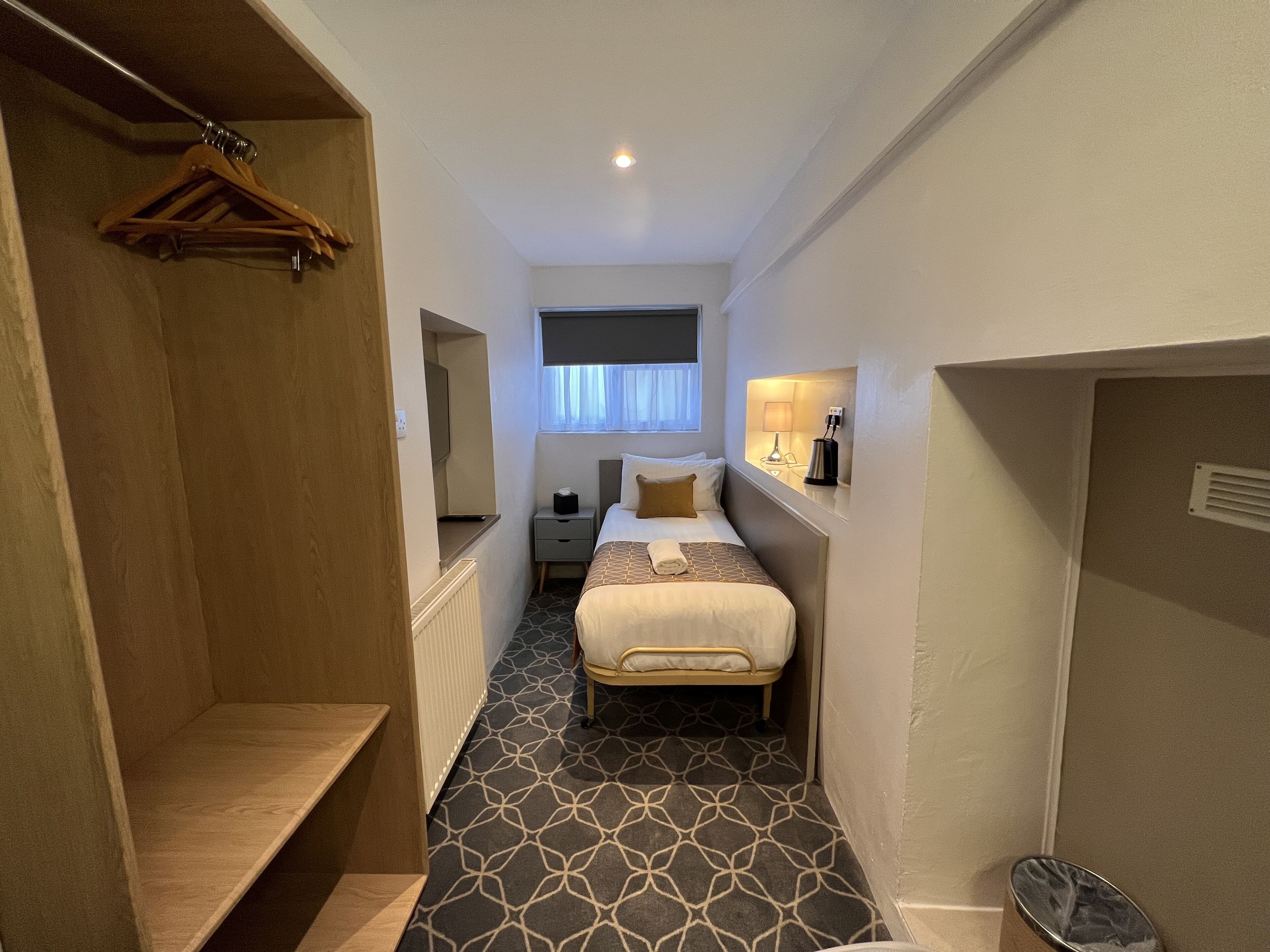 A narrow hotel room with a bed, bedside table, lamp, and a hanging coat rack.