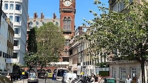 Un edificio histórico con una imponente torre de reloj, rodeado de arquitectura moderna y una animada calle.