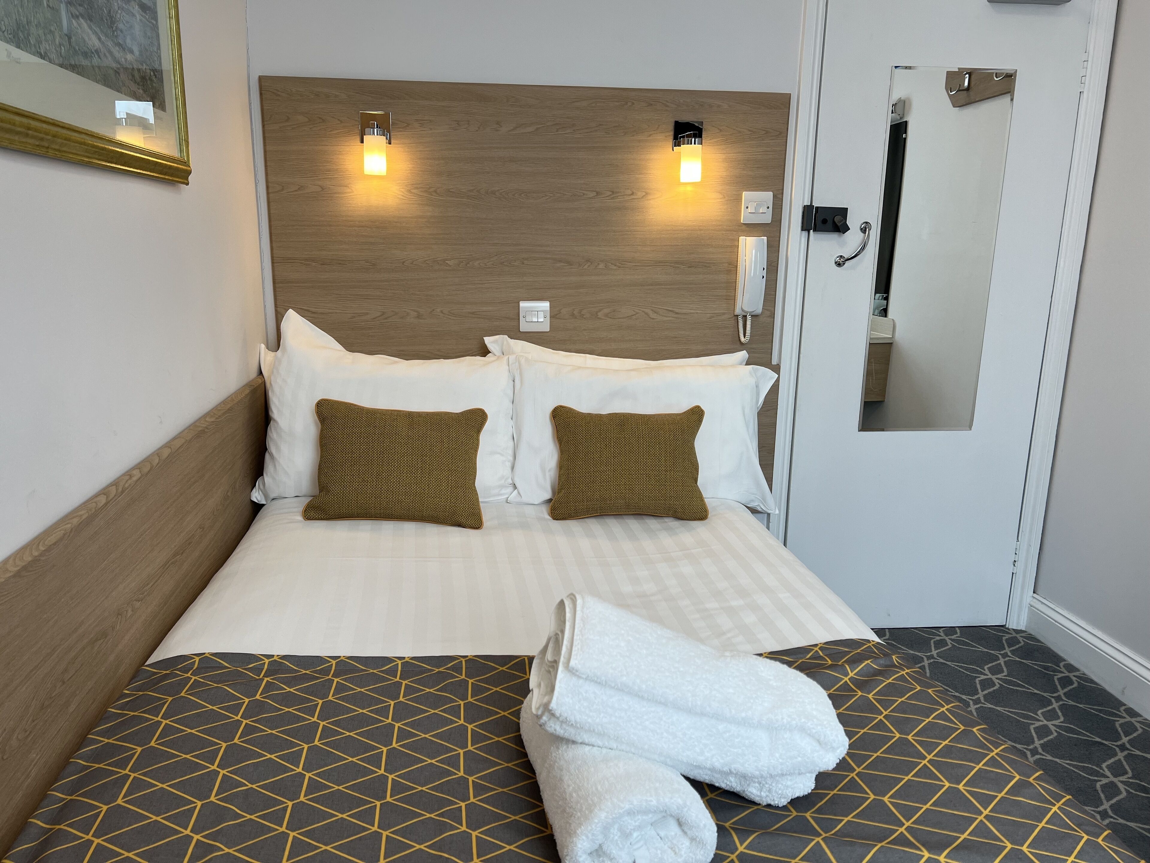 A hotel room with a wooden headboard, two white pillows, two brown pillows, a neatly made bed with a checkered bedspread, and a pair of folded towels on the floor.