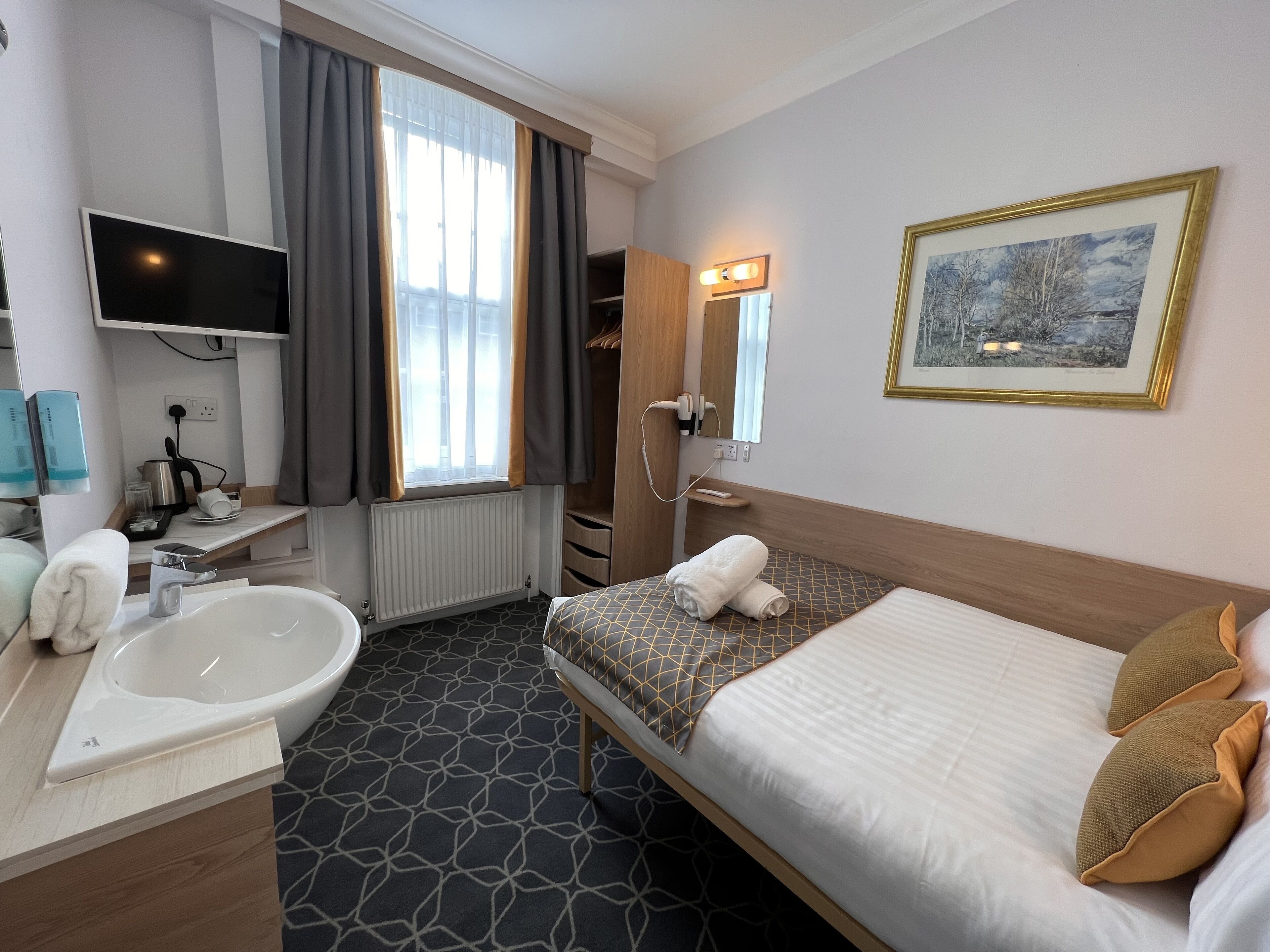 Ett hotellrum med en säng, ett stort fönster med gardiner, ett badkar, ett handfat, en spegel och en tv.