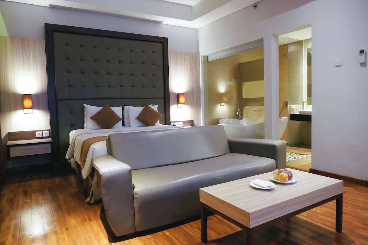 Una habitación de hotel moderna con una cama grande, un sofá gris, una mesa de centro de madera y un baño visible a través de una puerta abierta.