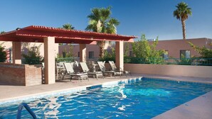 A swimming pool with lounge chairs and a covered area, surrounded by palm trees and a building. - GetAways at Havasu Dunes Resort (Lake Havasu City)