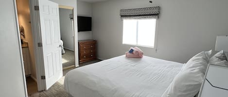 Une chambre à coucher comprenant un lit, un ventilateur de plafond, une fenêtre avec un lambrequin, une commode et une porte donnant sur une autre pièce.