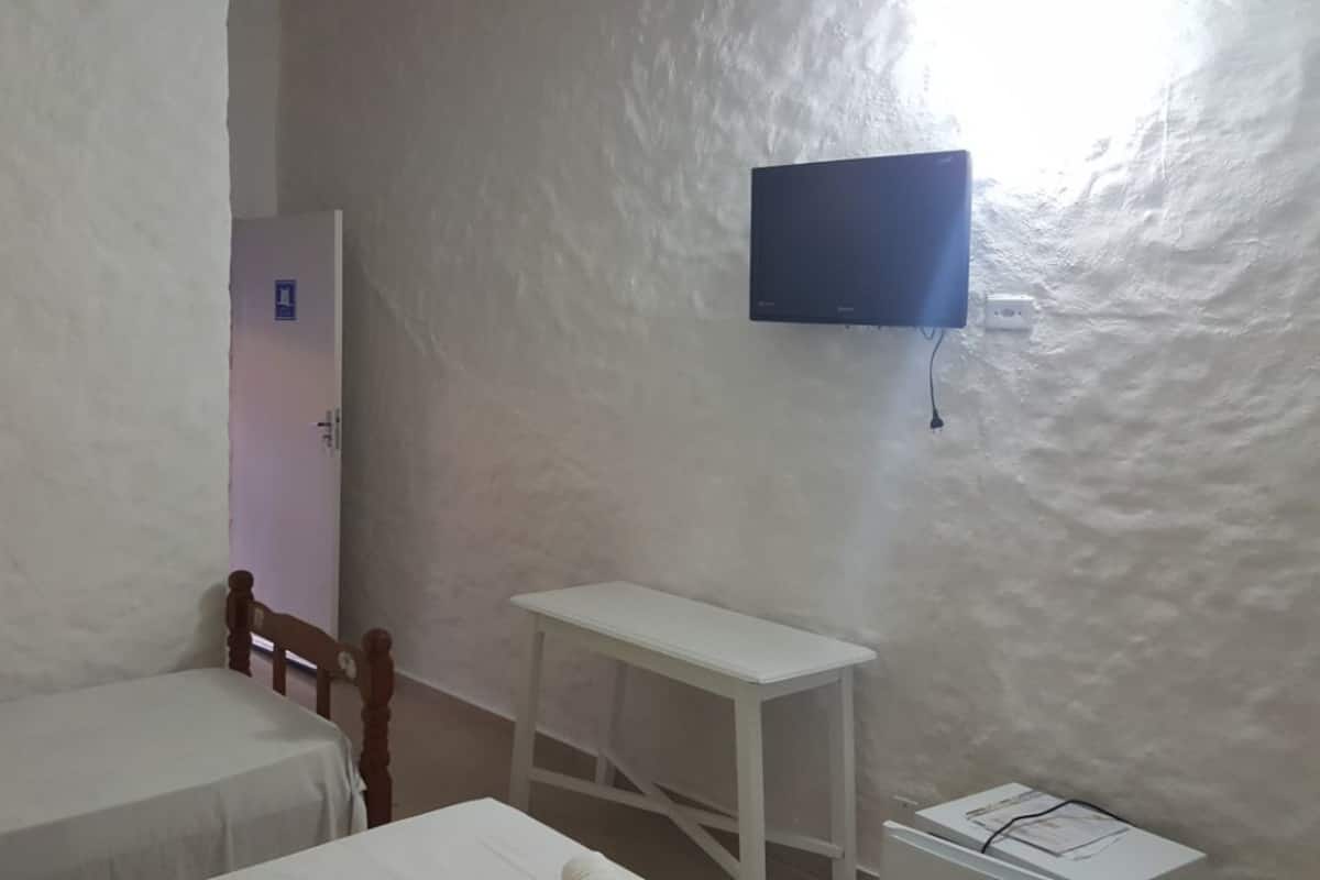 Ein kleines Zimmer mit einem Bett, einem Holzstuhl, einem weißen Tisch und einem an der Wand montierten Fernseher.