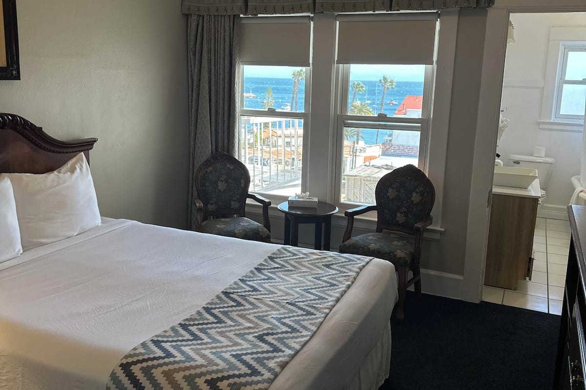 A hotel room with a bed, a desk, a chair, a window with a view of the ocean, and a ceiling fan.
