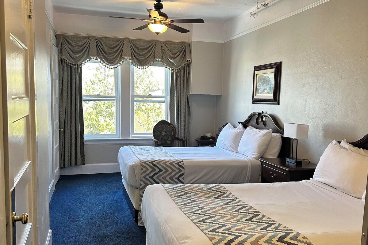 A hotel room with two beds, a ceiling fan, a window with curtains, a nightstand, and a framed picture on the wall.