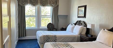 A hotel room with two beds, a ceiling fan, a window with curtains, a nightstand, and a framed picture on the wall.