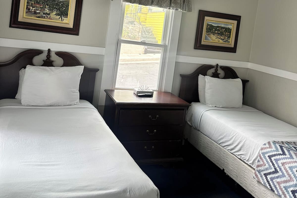 A hotel room with two beds, a ceiling fan, a window with a view, and framed pictures on the walls.