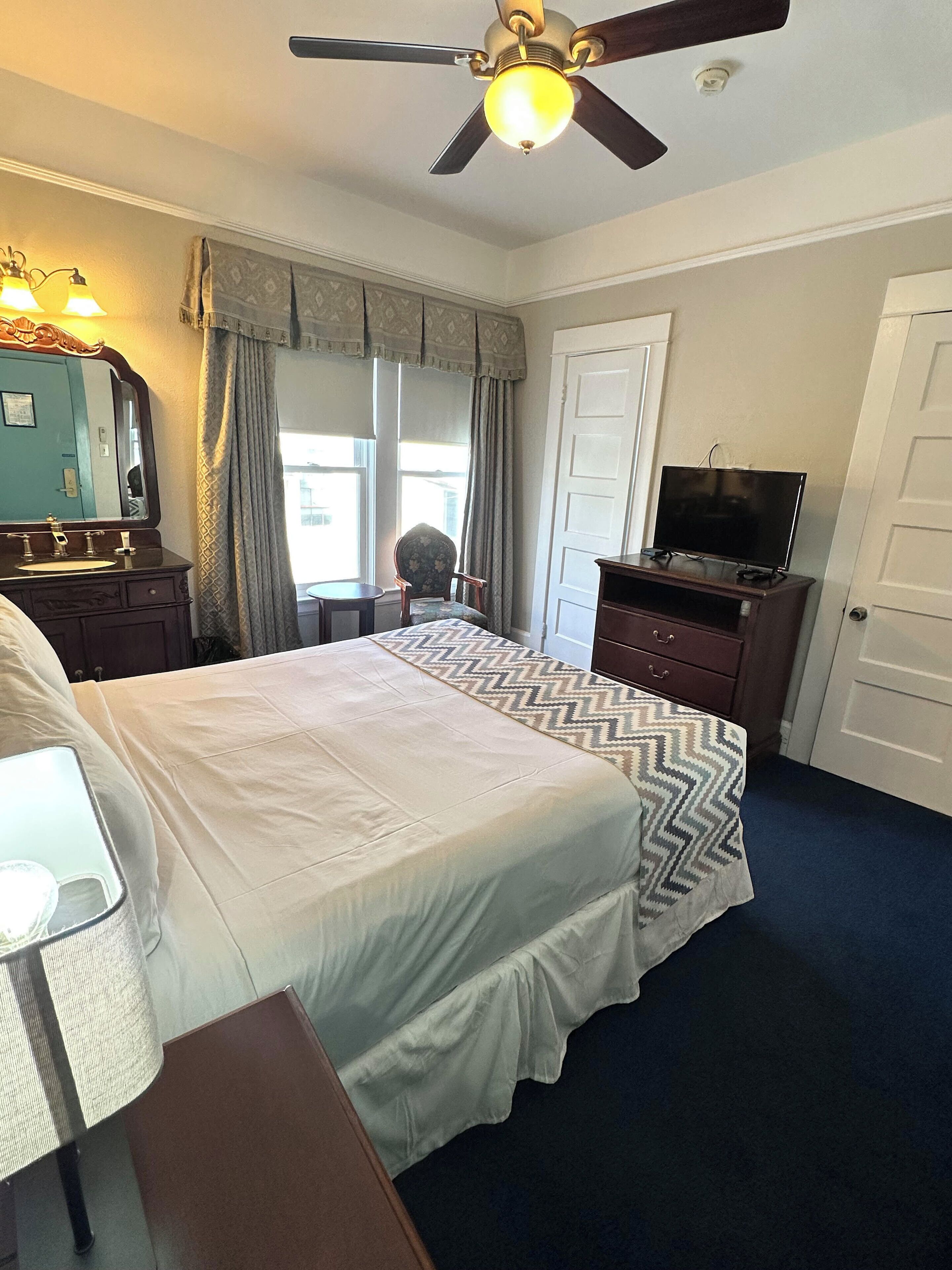 A hotel room with a bed, a desk, a chair, a television, and a ceiling fan.