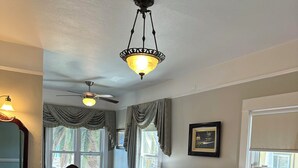 A hotel room with two beds, a ceiling fan, a chandelier, a desk with a lamp, a chair, and a window with curtains. - Studio 9 Hotel Catalina (Avalon)