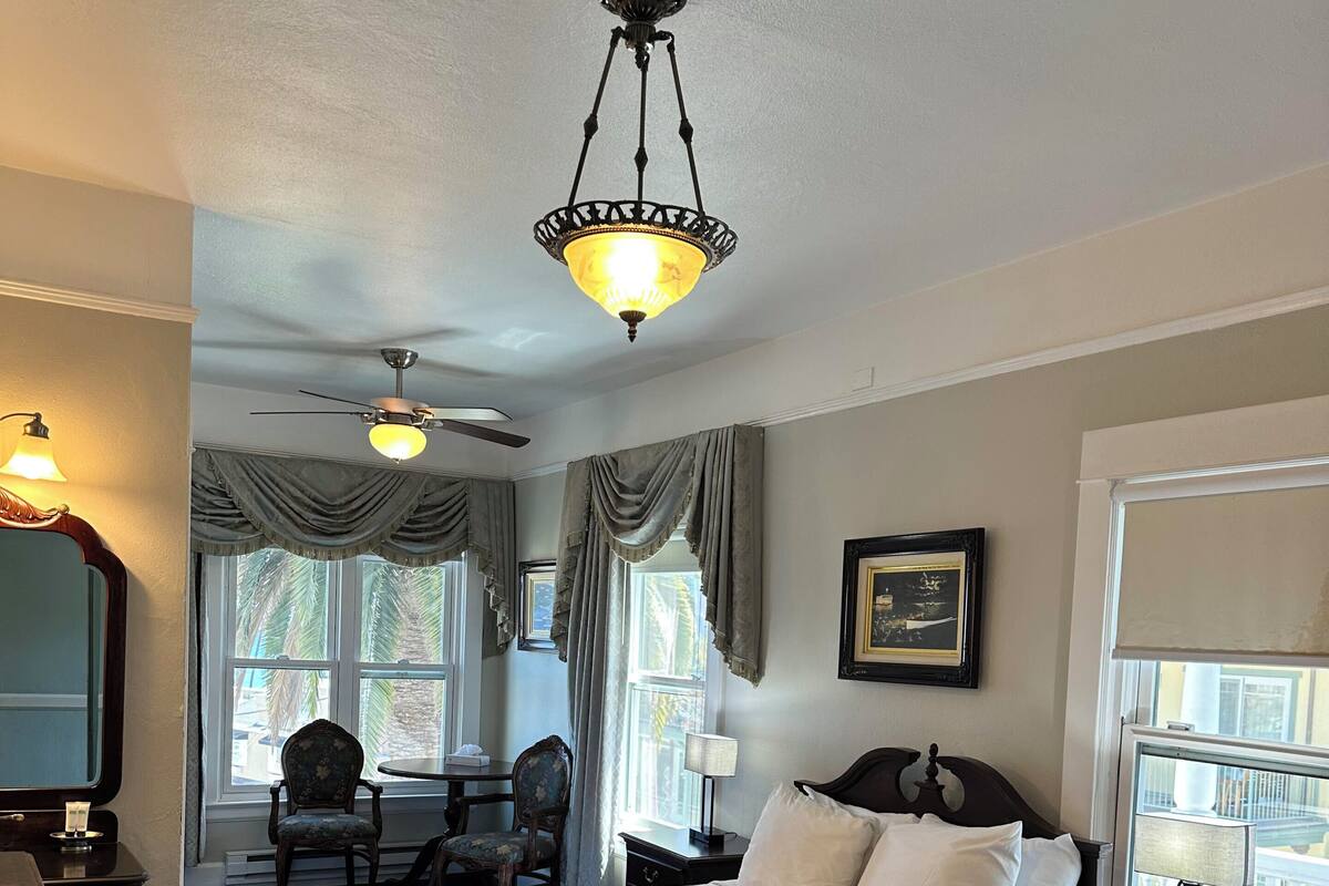 A hotel room with two beds, a ceiling fan, a chandelier, a desk with a lamp, a chair, and a window with curtains.