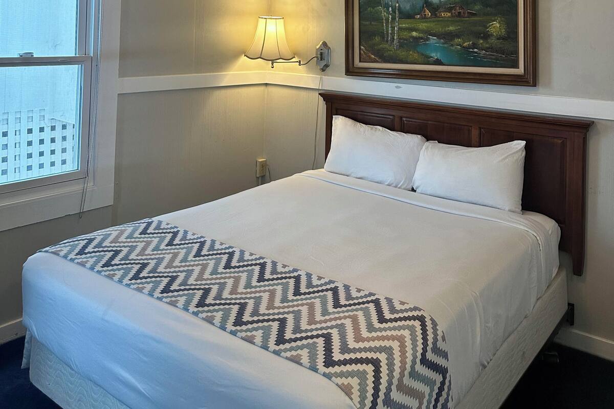 A hotel room with a bed, a ceiling fan, a painting, and a window.