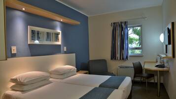 Une chambre d’hôtel avec deux lits, un bureau, une chaise, une fenêtre avec des rideaux et un téléviseur fixé au mur.