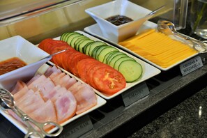 Noutopöydässä on erilliset osiot, joissa on selkeät kyltit: kinkku, tomaatit, kurkut, cheddar- ja mozzarella-juusto sekä pieni kulho kastiketta.
