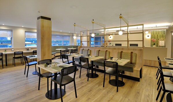 A modern restaurant with wooden flooring, black chairs, and tables. There are large windows providing natural light and views of the outside.
