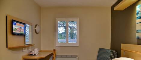 A hotel room with a bed, a desk with a chair, a television, a window with blinds, and a wall clock.