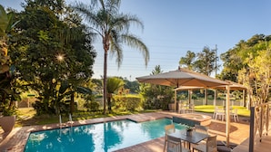 Una piscina rectangular con borde de azulejos, rodeada por un patio de ladrillo. Hay una zona de estar al aire libre cubierta con mesas y sillas.