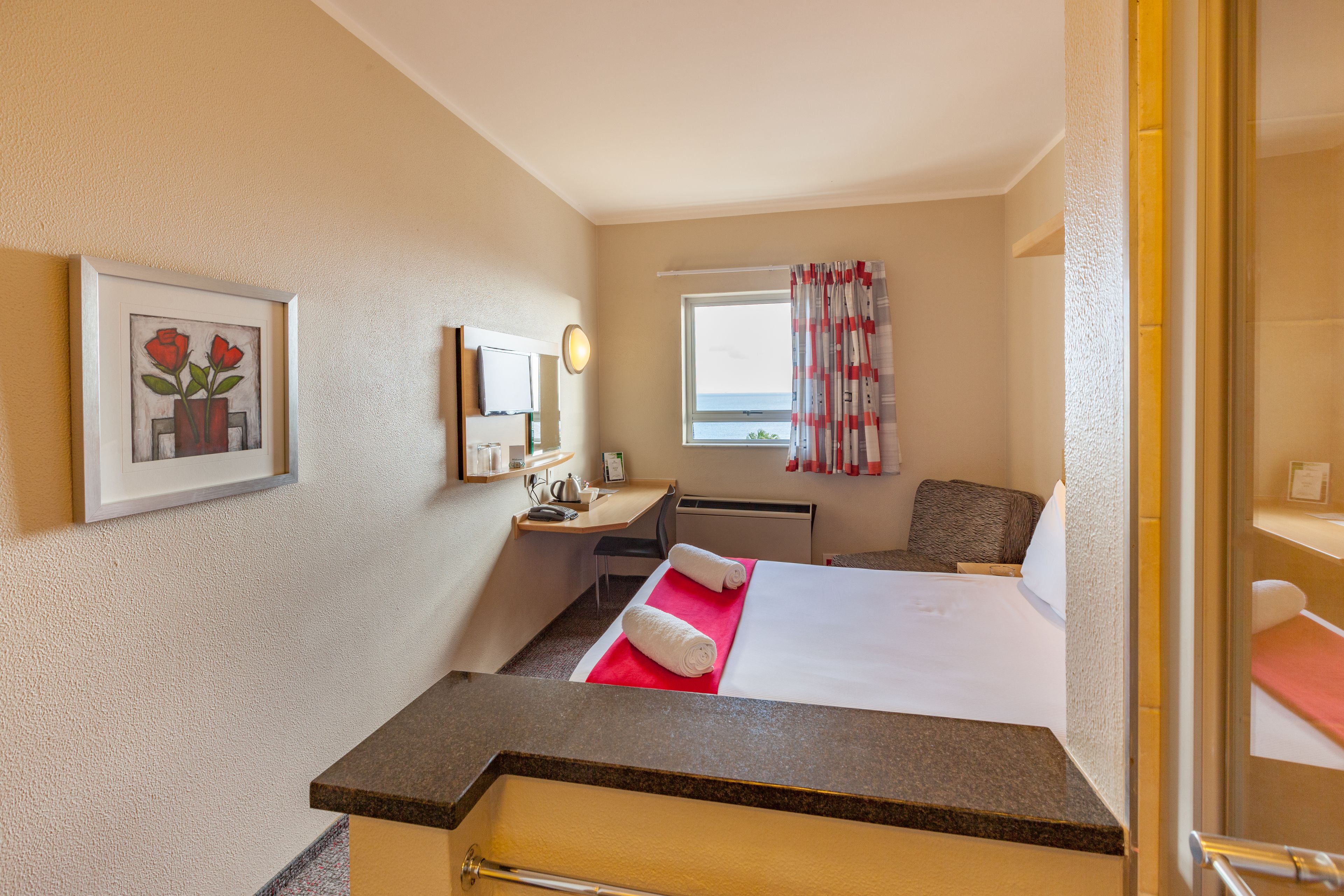 Une chambre d’hôtel avec un lit, un bureau, une chaise, une fenêtre avec des rideaux, un tableau encadré au mur et une porte de salle de bain.