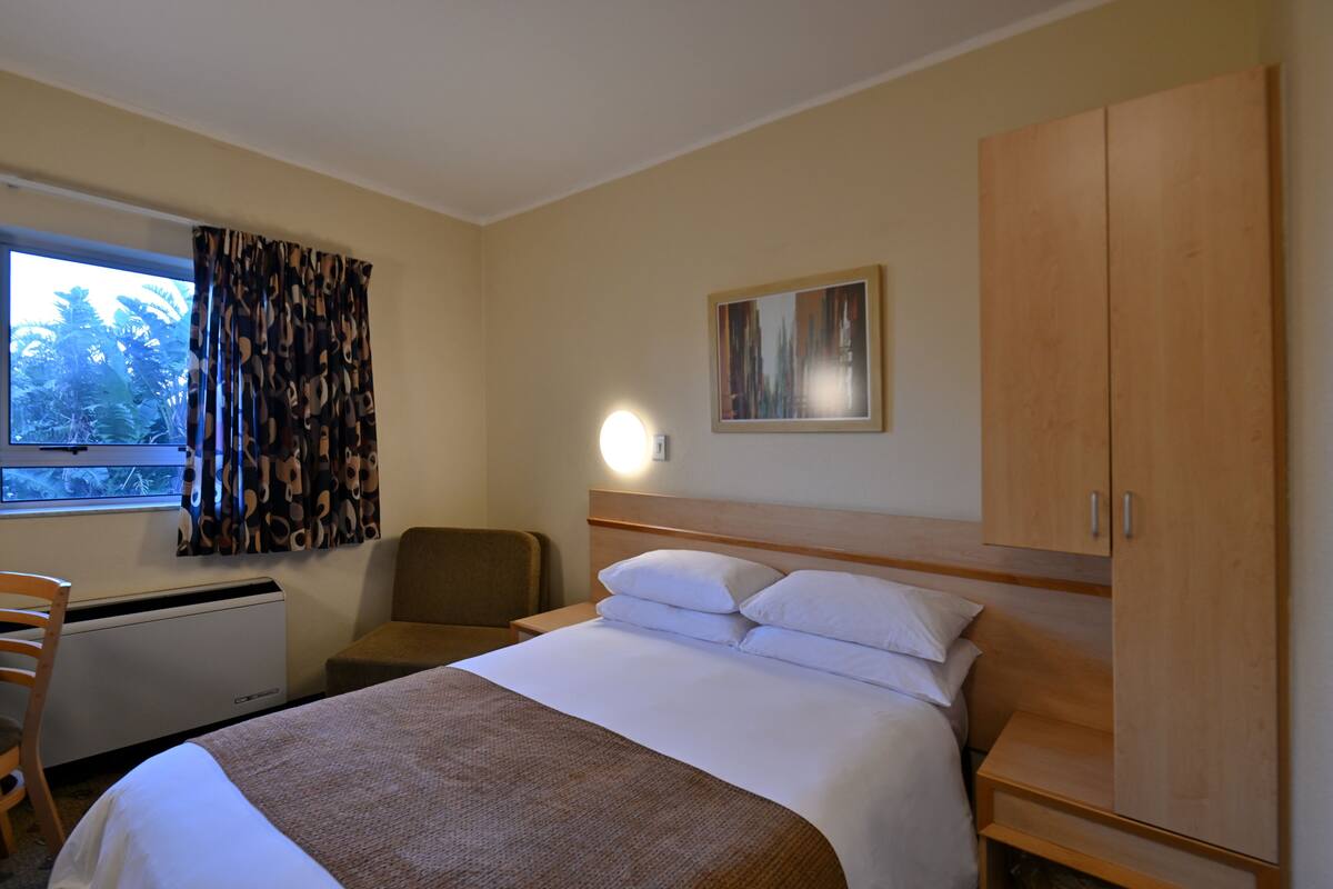 Hotelkamer met een bed, een stoel, een bureau, een kledingkast, een raam met gordijnen en een wandlamp.
