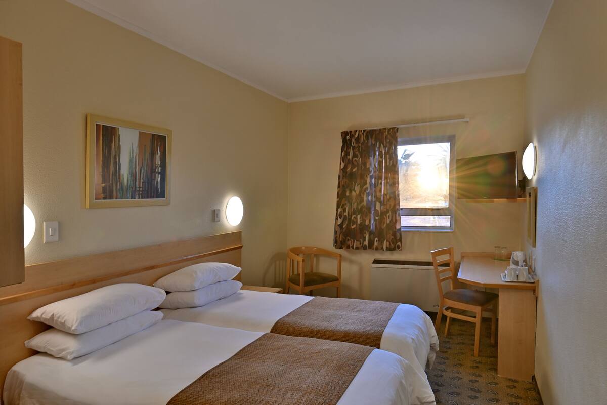 Een hotelkamer met twee bedden, een bureau, een stoel, een raam met gordijnen, een schilderij aan de muur en een ronde spiegel.