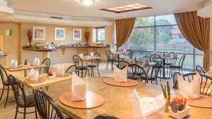Une salle à manger d’hôtel avec de nombreuses tables et chaises, un comptoir où sont exposés différents plats, et de grandes fenêtres dotées de rideaux.