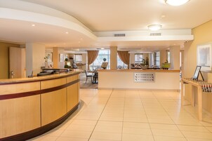 Zona de recepción de hotel con un mostrador curvo, un área de estar con sillas y un cartel con los números de habitaciones e indicaciones.