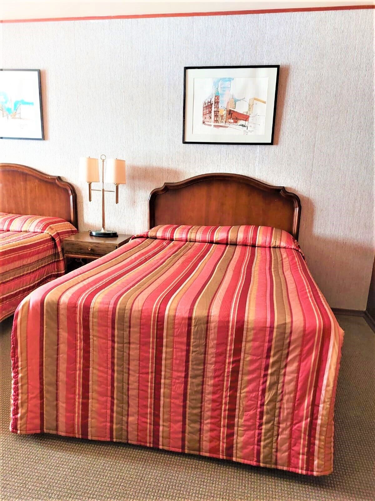 A hotel room with two beds, each with a striped red and beige bedspread. There is a nightstand with a lamp between the beds.