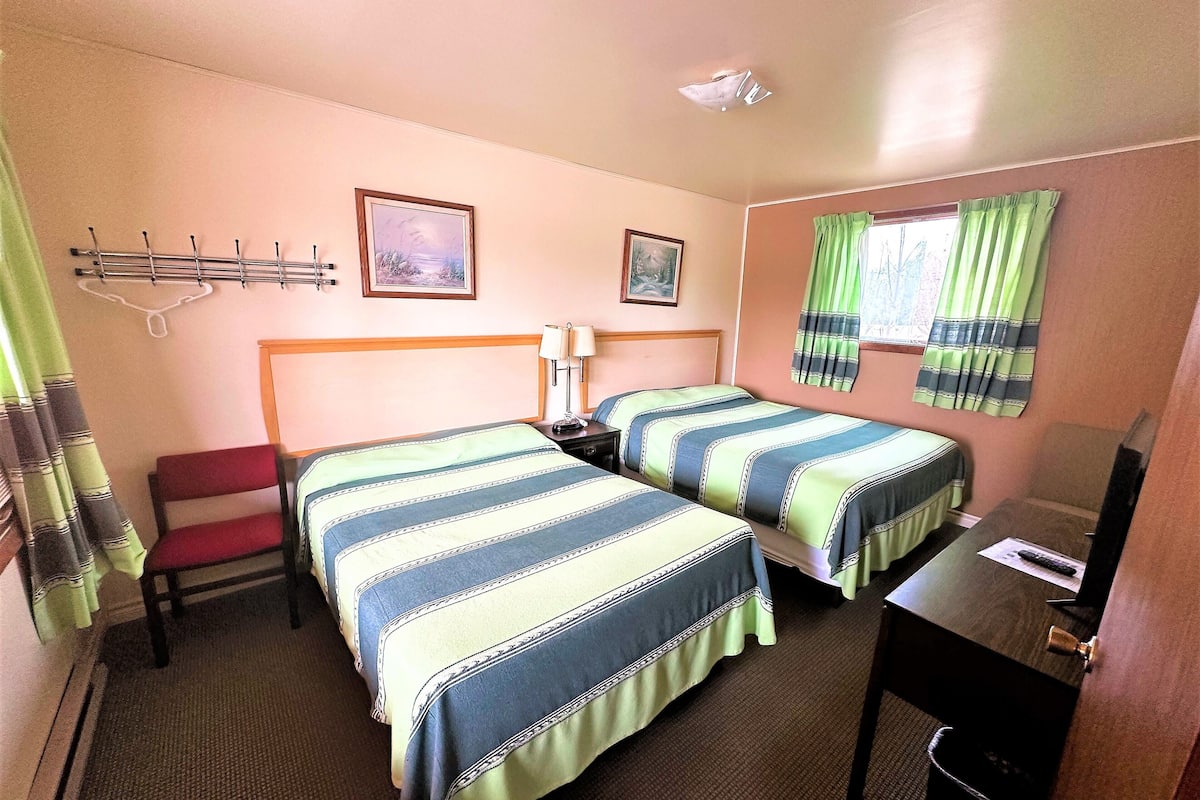 A hotel room with two beds, a desk, a chair, a small table, a lamp, a picture frame, a window with curtains, and a ceiling light.