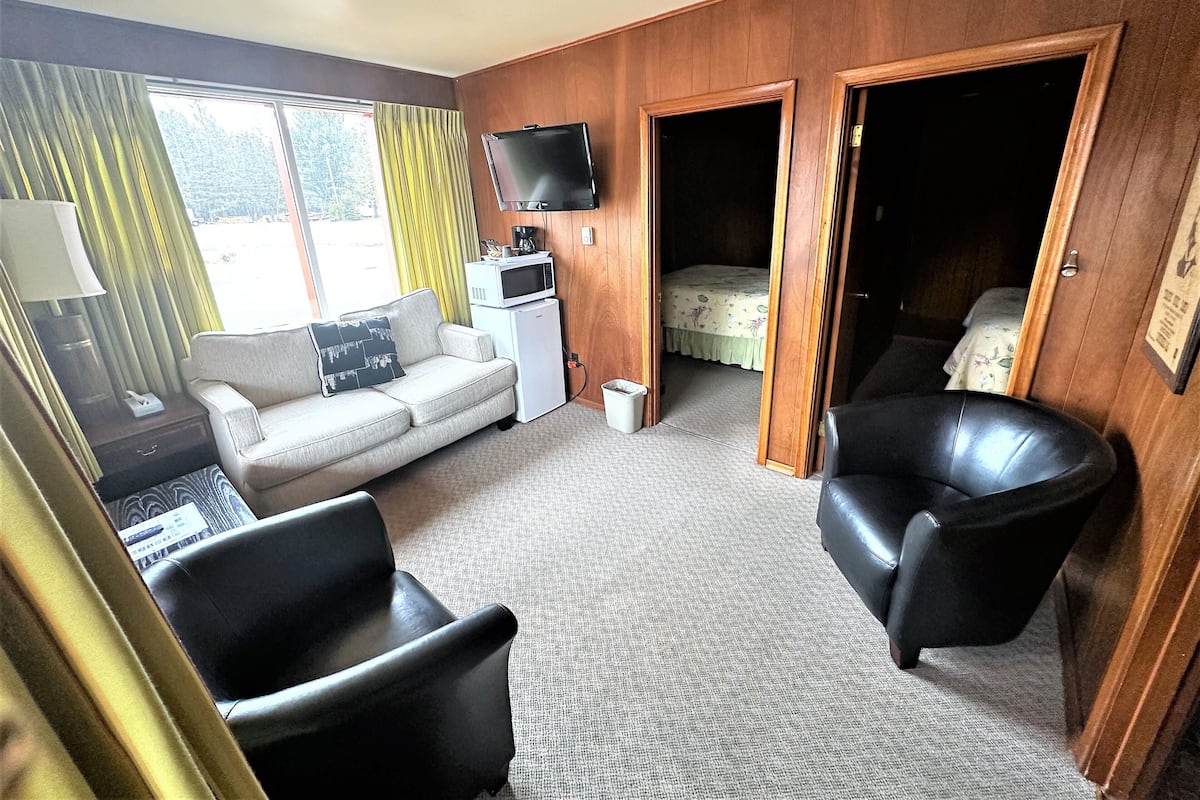 A compact room with a sofa, a black leather chair, a small white fridge, a microwave, and a bed with a floral bedspread.