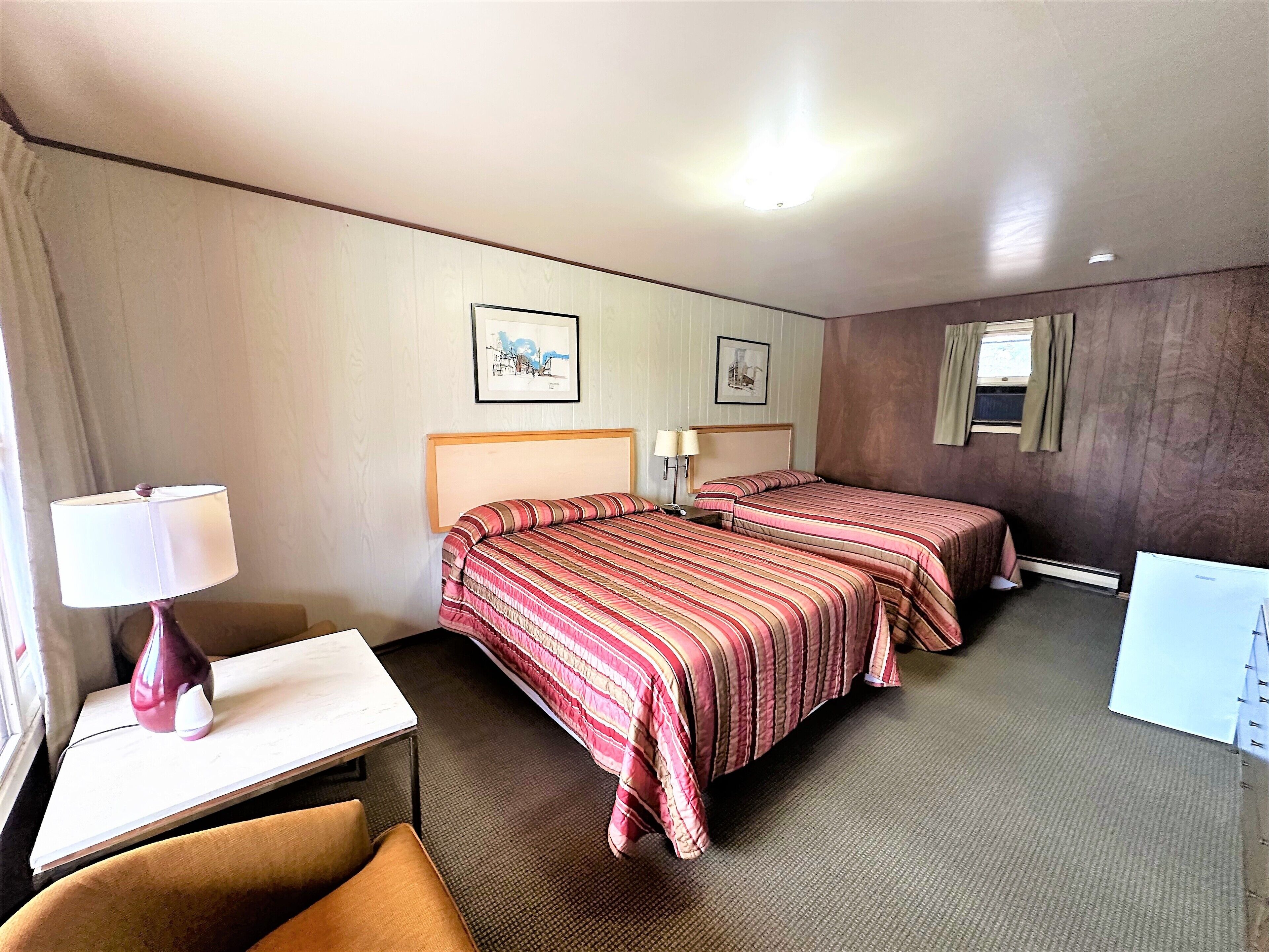 A hotel room with two beds, a desk, a chair, a lamp, and two framed pictures on the wall.