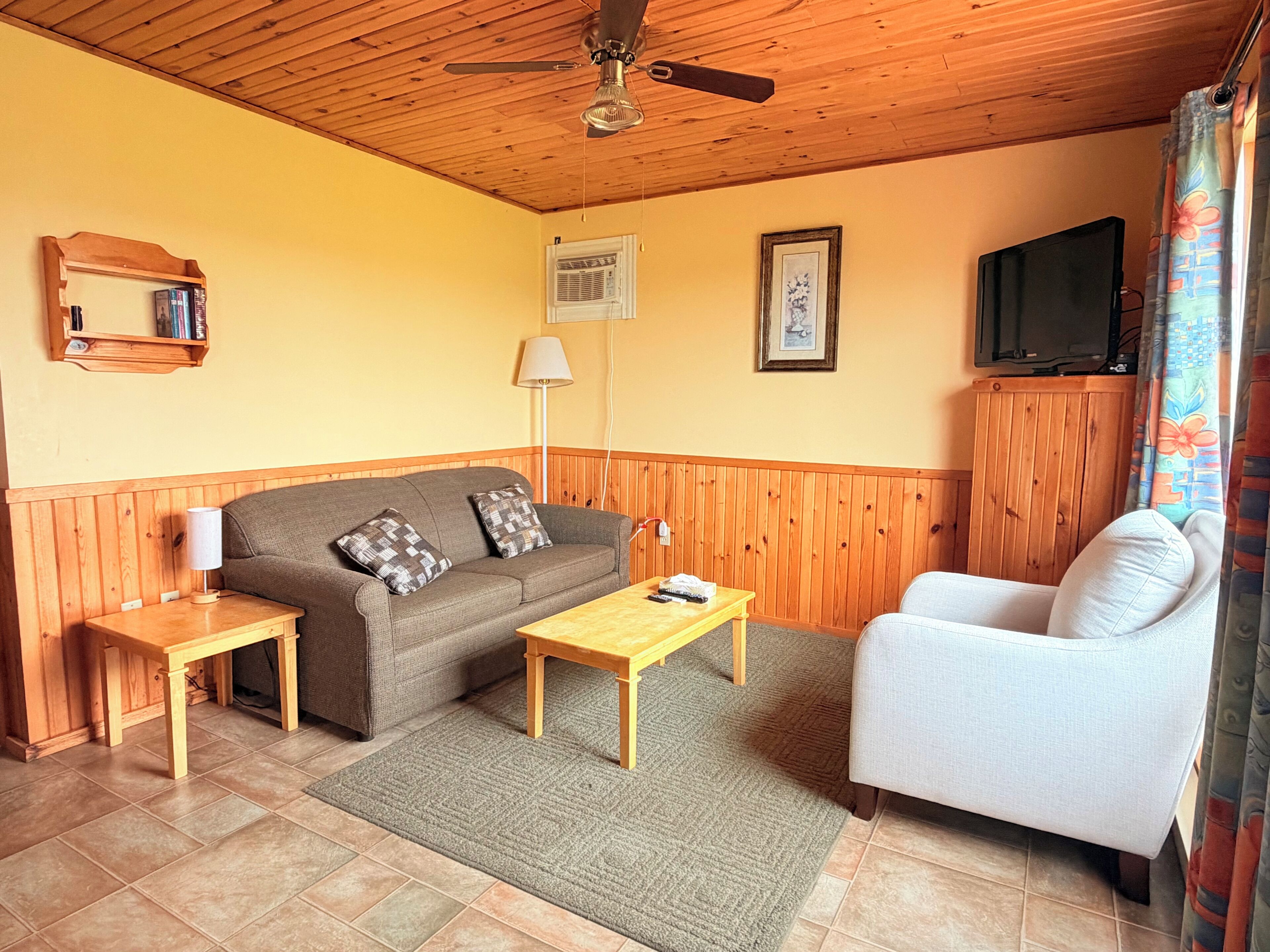 A room with a wooden ceiling, a sofa, a chair, a small table, a TV, and a window with curtains.
