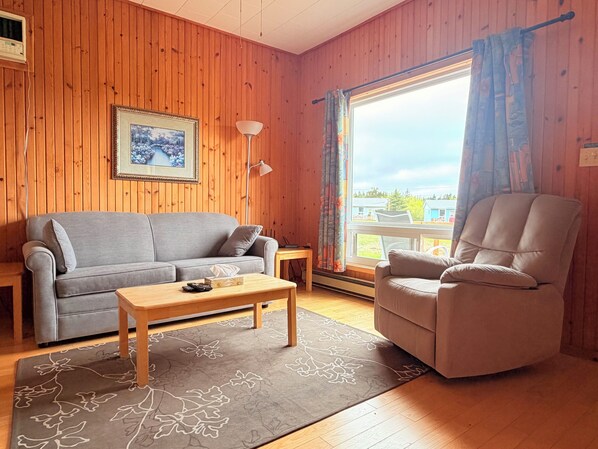 A wooden-paneled room with a grey sofa, a wooden coffee table, a beige armchair, a framed picture on the wall, and a window with curtains.