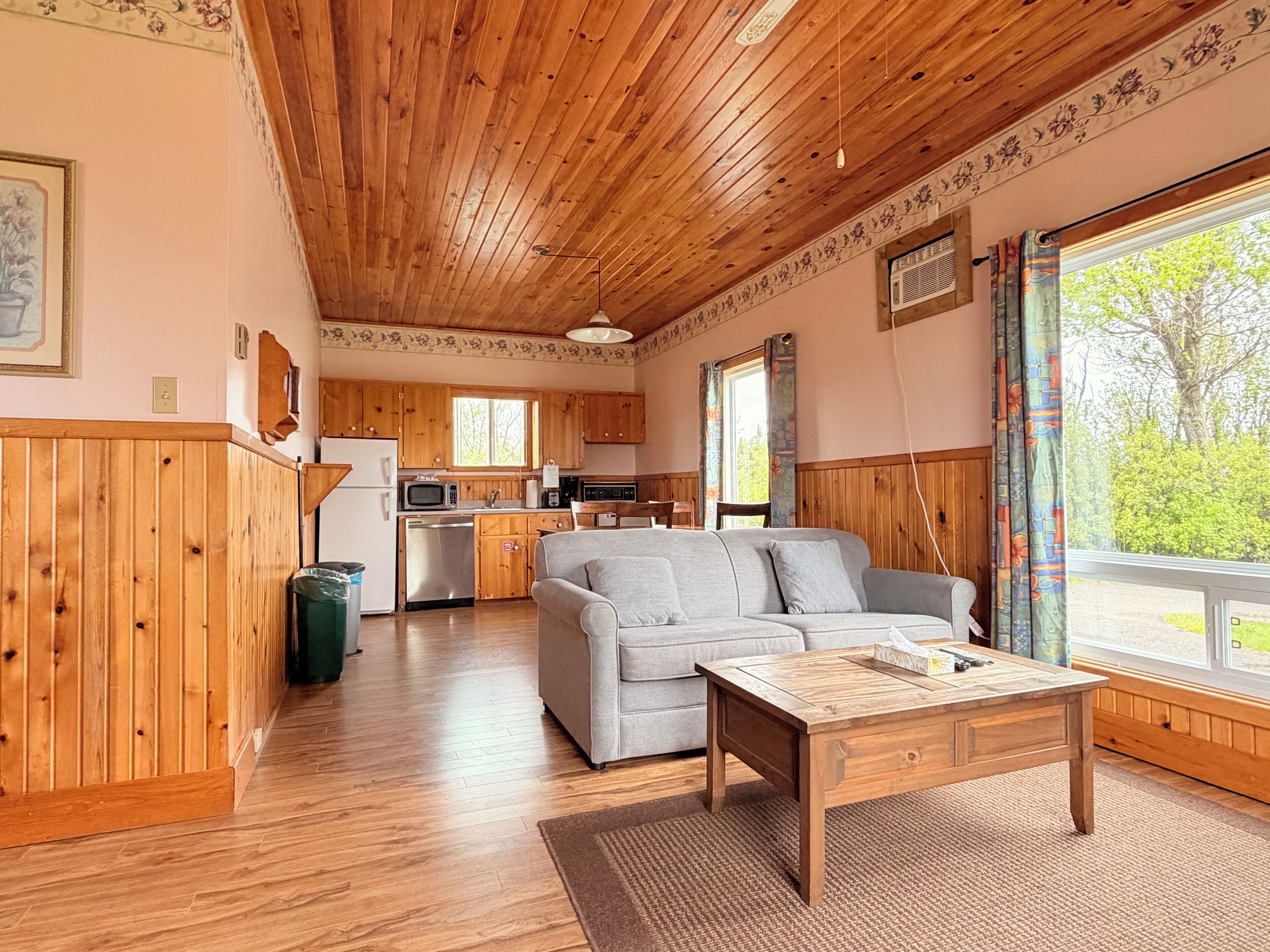 L’intérieur d’un chalet accueillant, avec un plafond en bois, un coin cuisine, un canapé gris et une grande fenêtre agrémentée de rideaux.