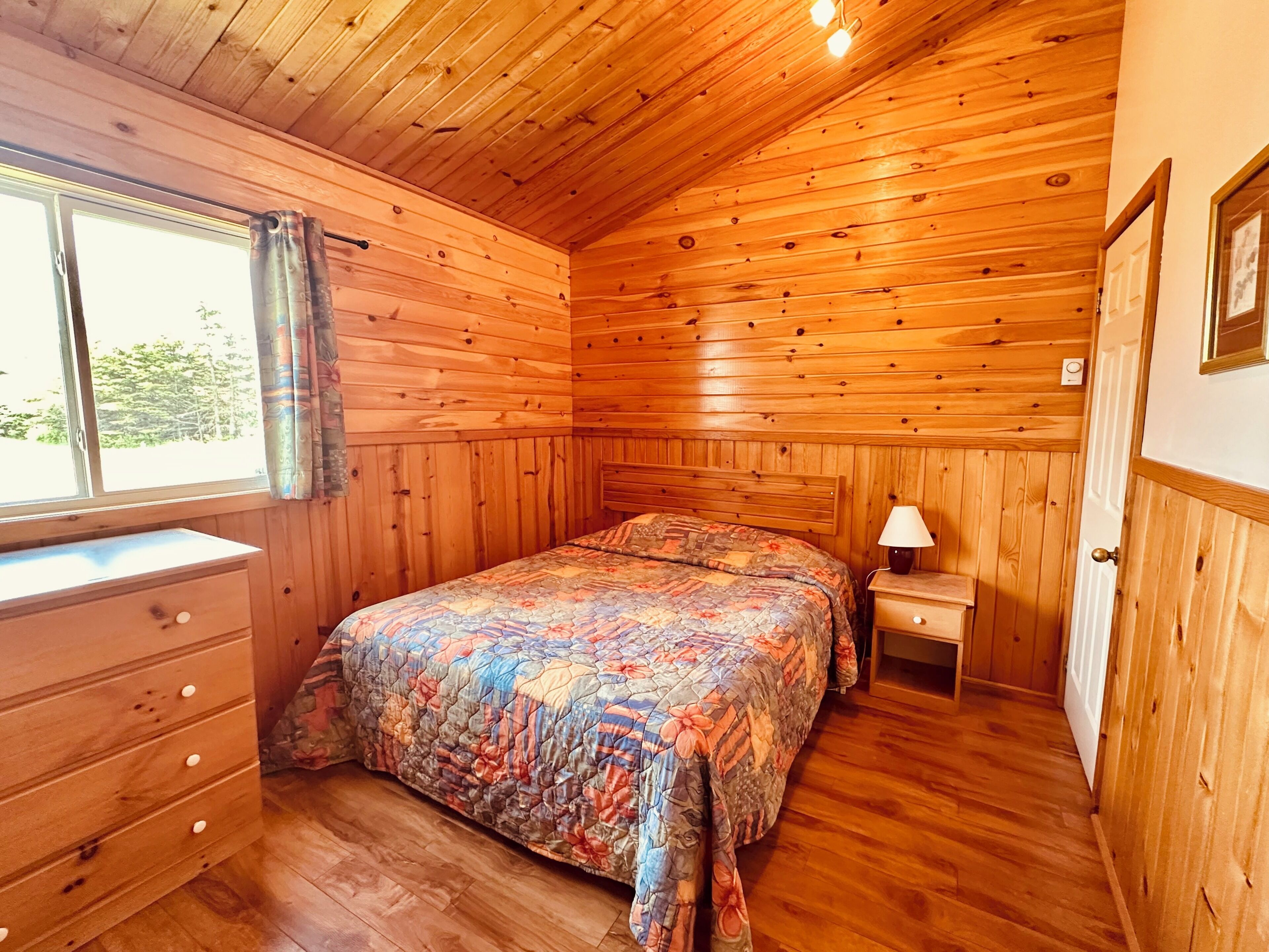 A wooden-paneled bedroom with a bed, dresser, bedside table, and window.