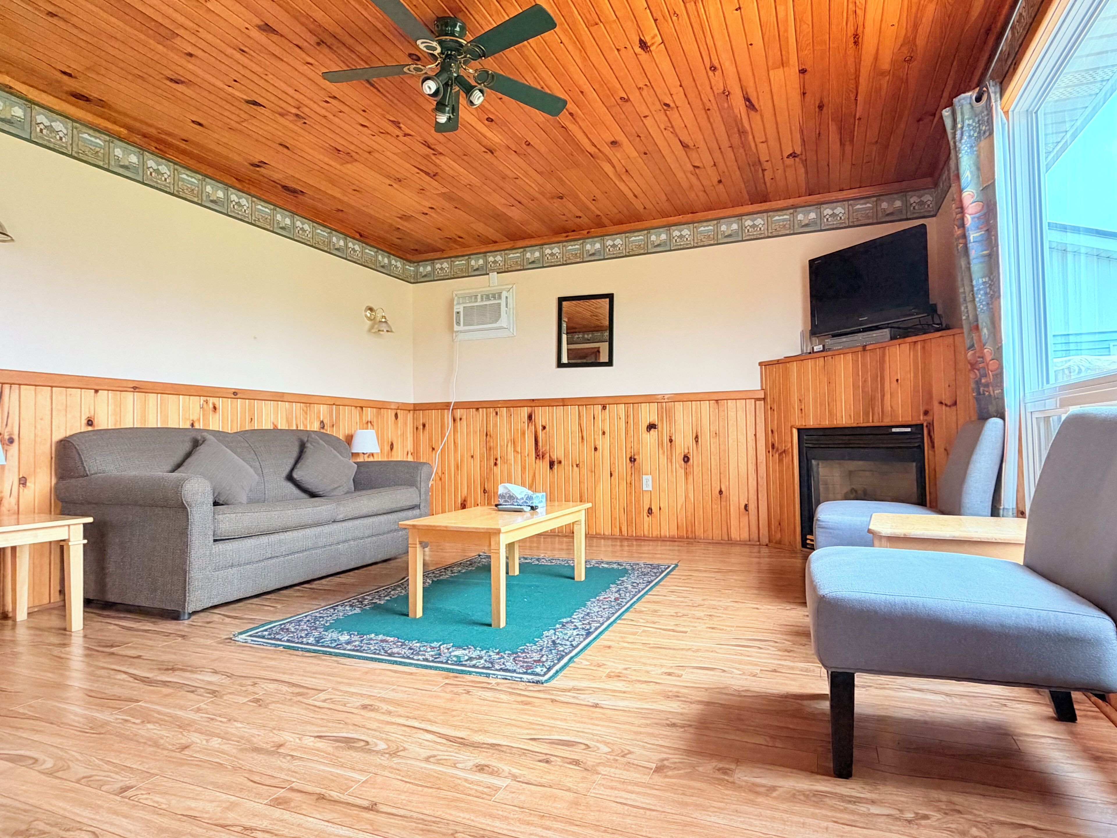 A room with a wooden ceiling, a ceiling fan, a fireplace, a television, a sofa, a chair, and a small table.