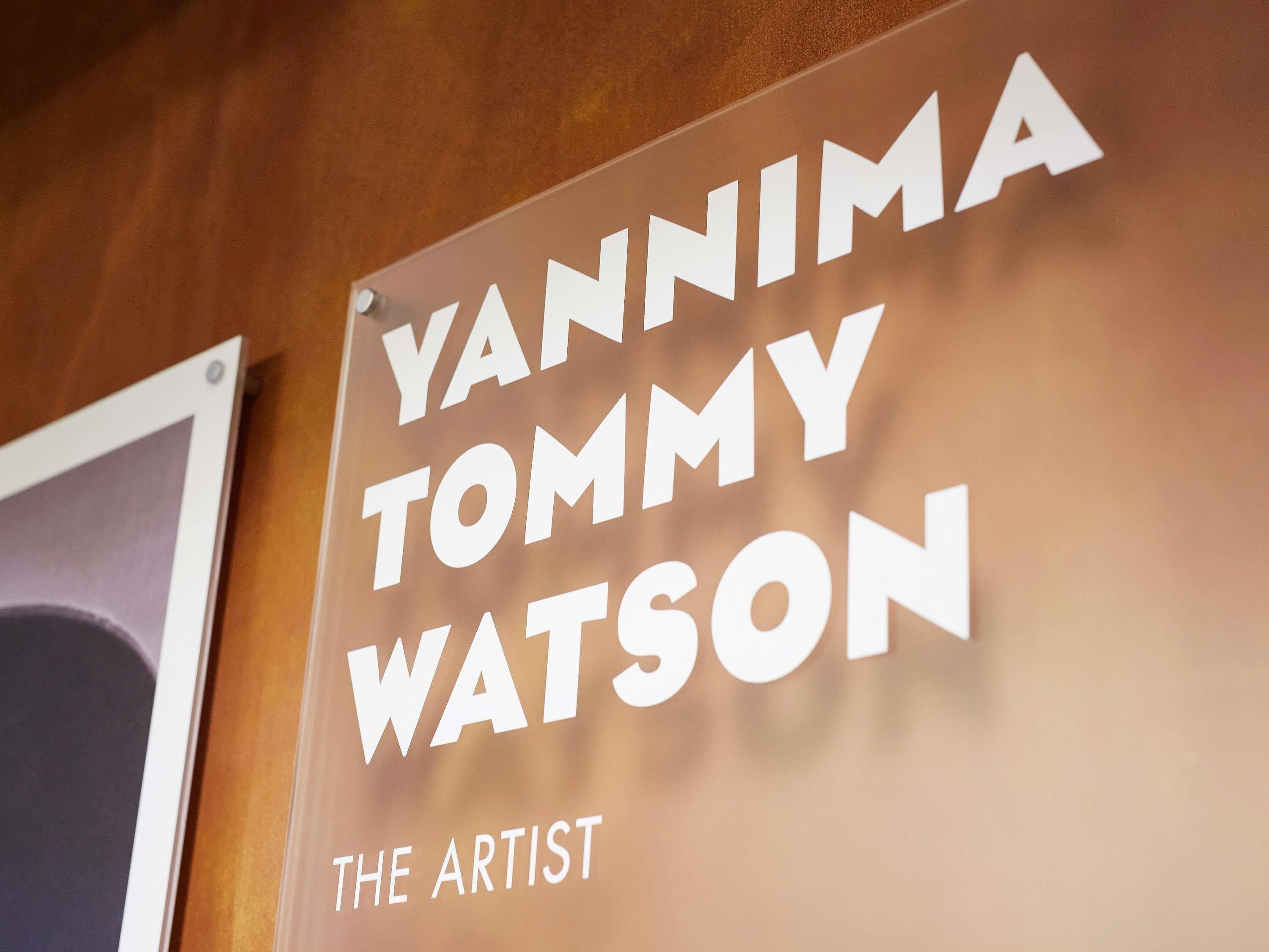 Un letrero con el texto 'YANNIMA TOMMY WATSON THE ARTIST' en una pared de madera.