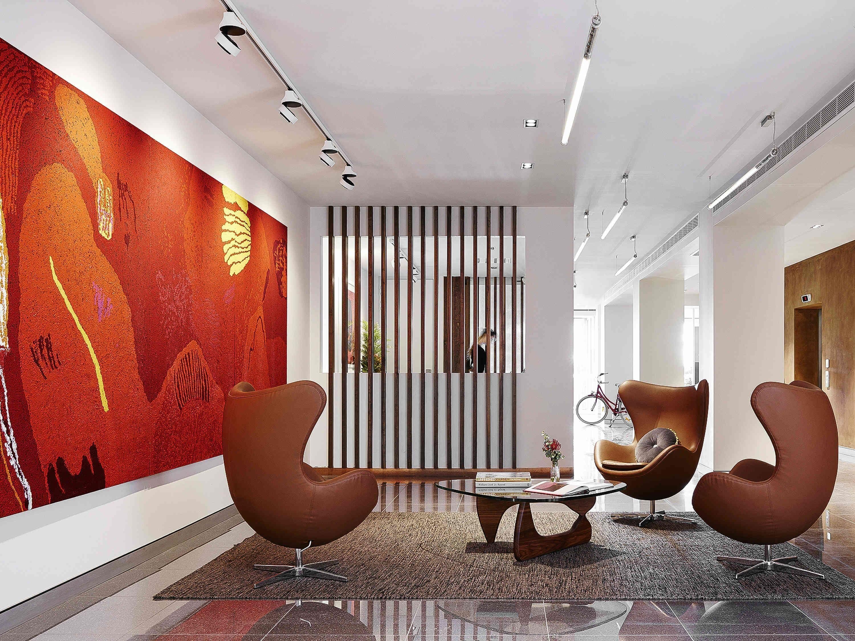 Un vestíbulo moderno con un gran mural abstracto, dos sillones marrones, una mesa de centro de vidrio y un separador a rayas.