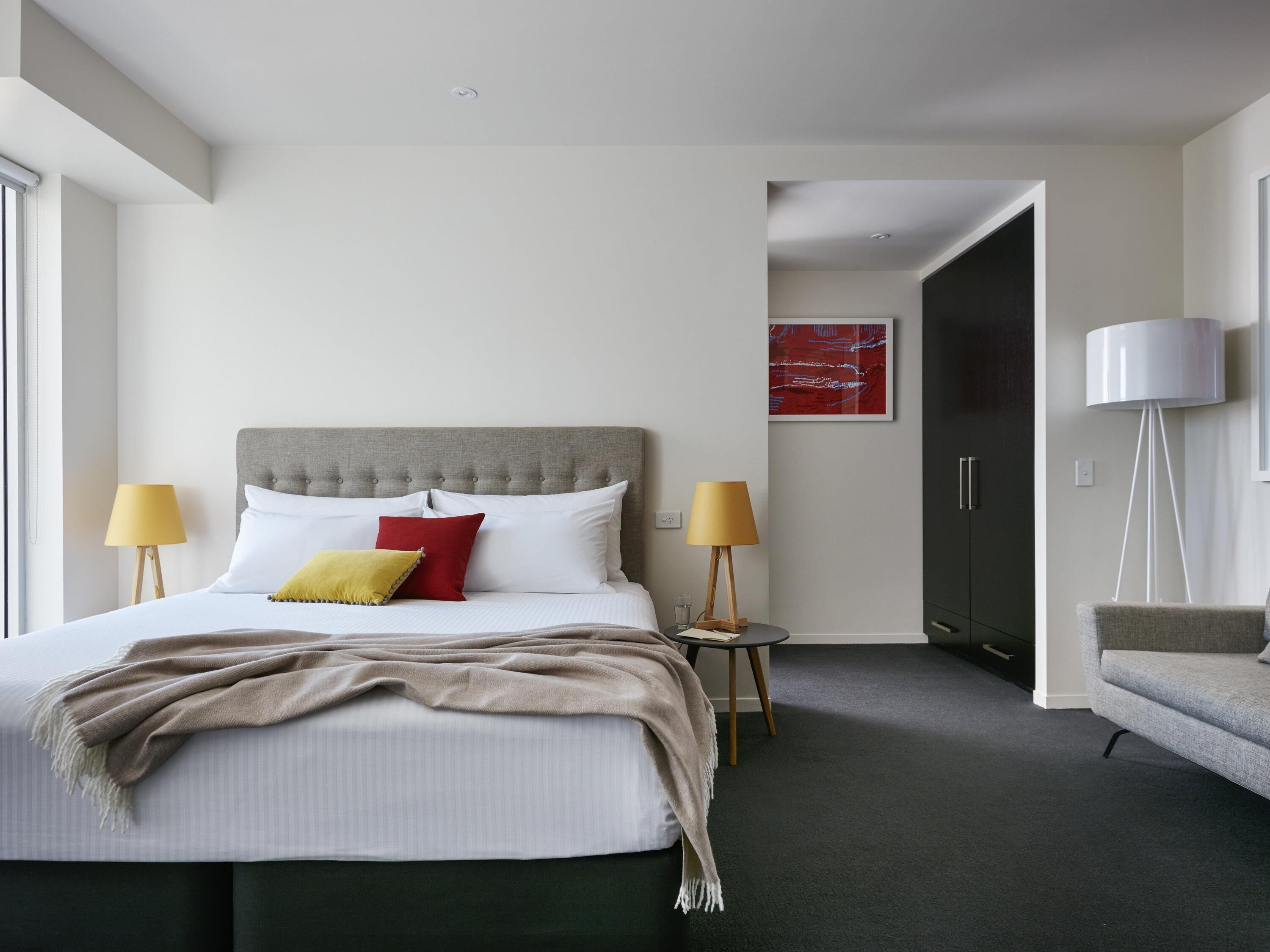 Una habitación de hotel moderna con cama, lámparas de noche, mesitas de noche, una silla y un cuadro en la pared.