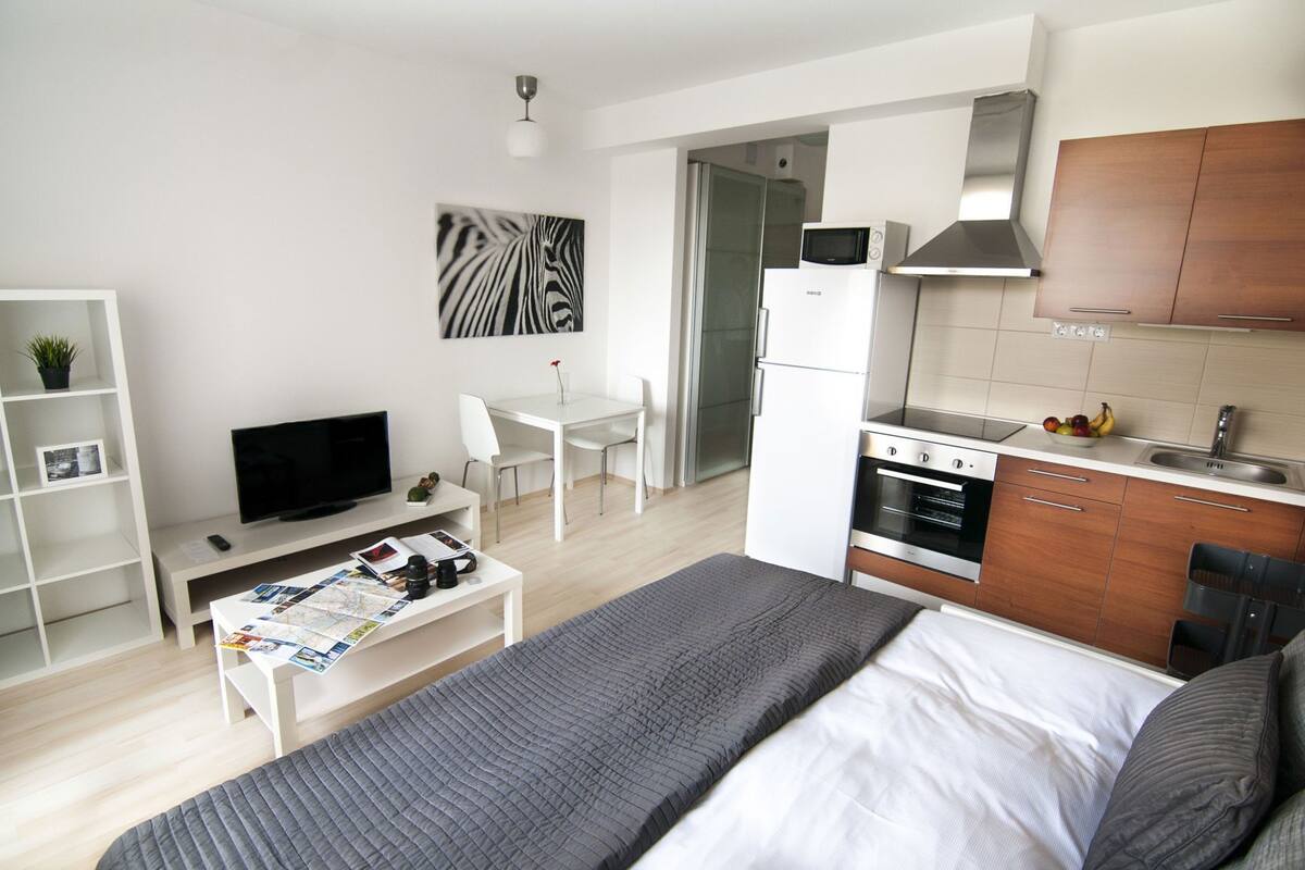 A modern hotel room with a bed, a desk, a TV, a kitchenette, and a dining area.