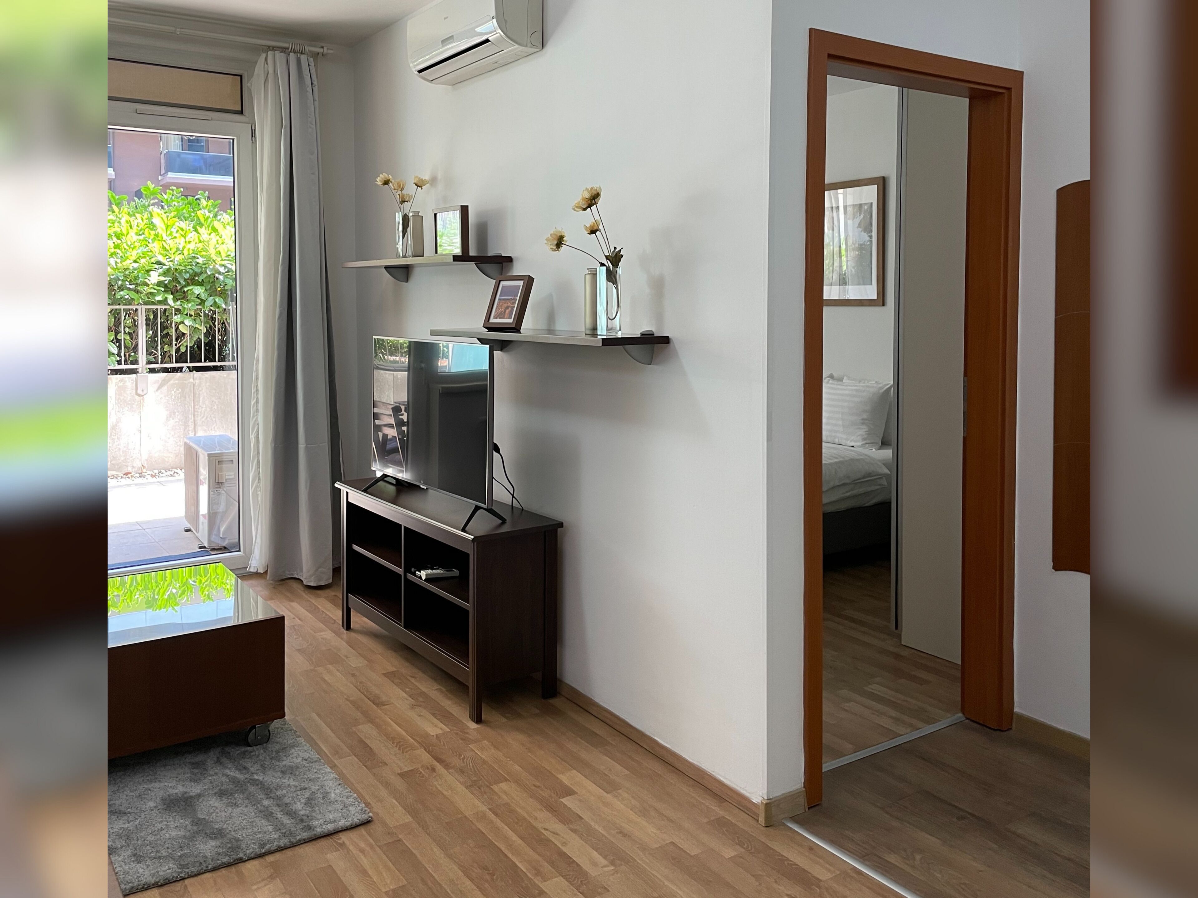 A modern living room with a flat-screen TV, wooden flooring, a small entertainment unit, a mirror, and a door leading to another room.
