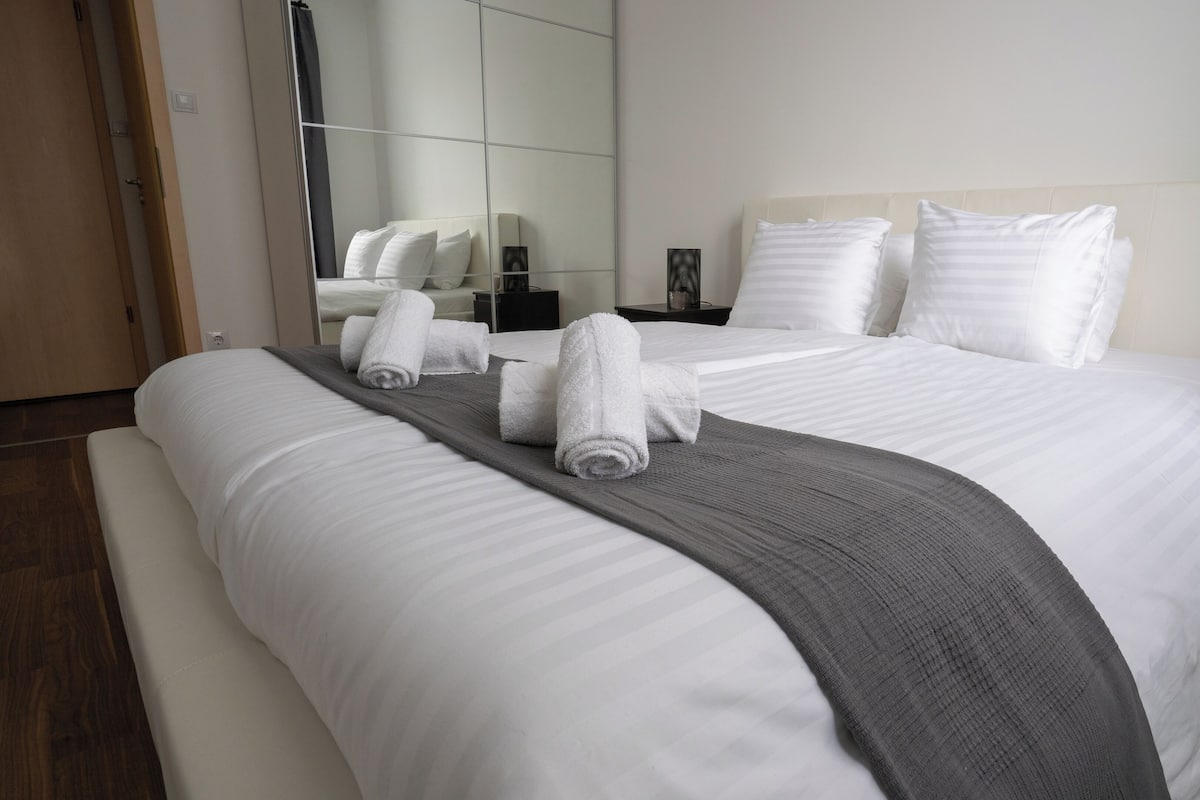 A hotel room with a large bed, a grey throw blanket, and neatly folded towels.