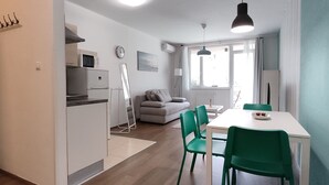 Une cuisine moderne avec des meubles blancs, un four à micro-ondes et un réfrigérateur. On y trouve une table à manger avec des chaises vertes et une nappe blanche. Un canapé gris se trouve au fond de la pièce, et une fenêtre laisse entrer la lumière naturelle.