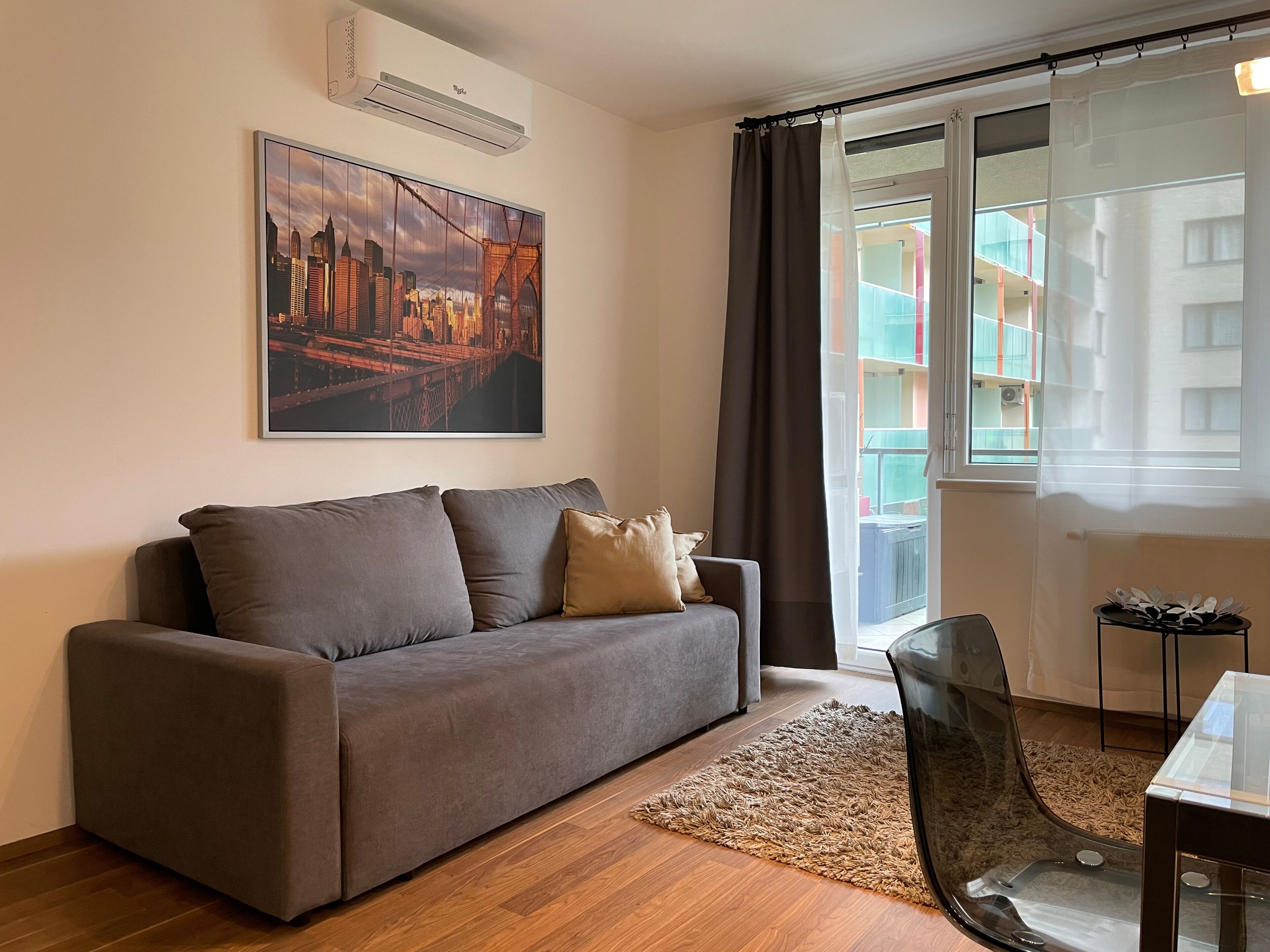 Een moderne woonkamer met een grijze bank, een stadsgezicht schilderij, een glazen tafel, en uitzicht op een balkon met kleurrijke gebouwen.